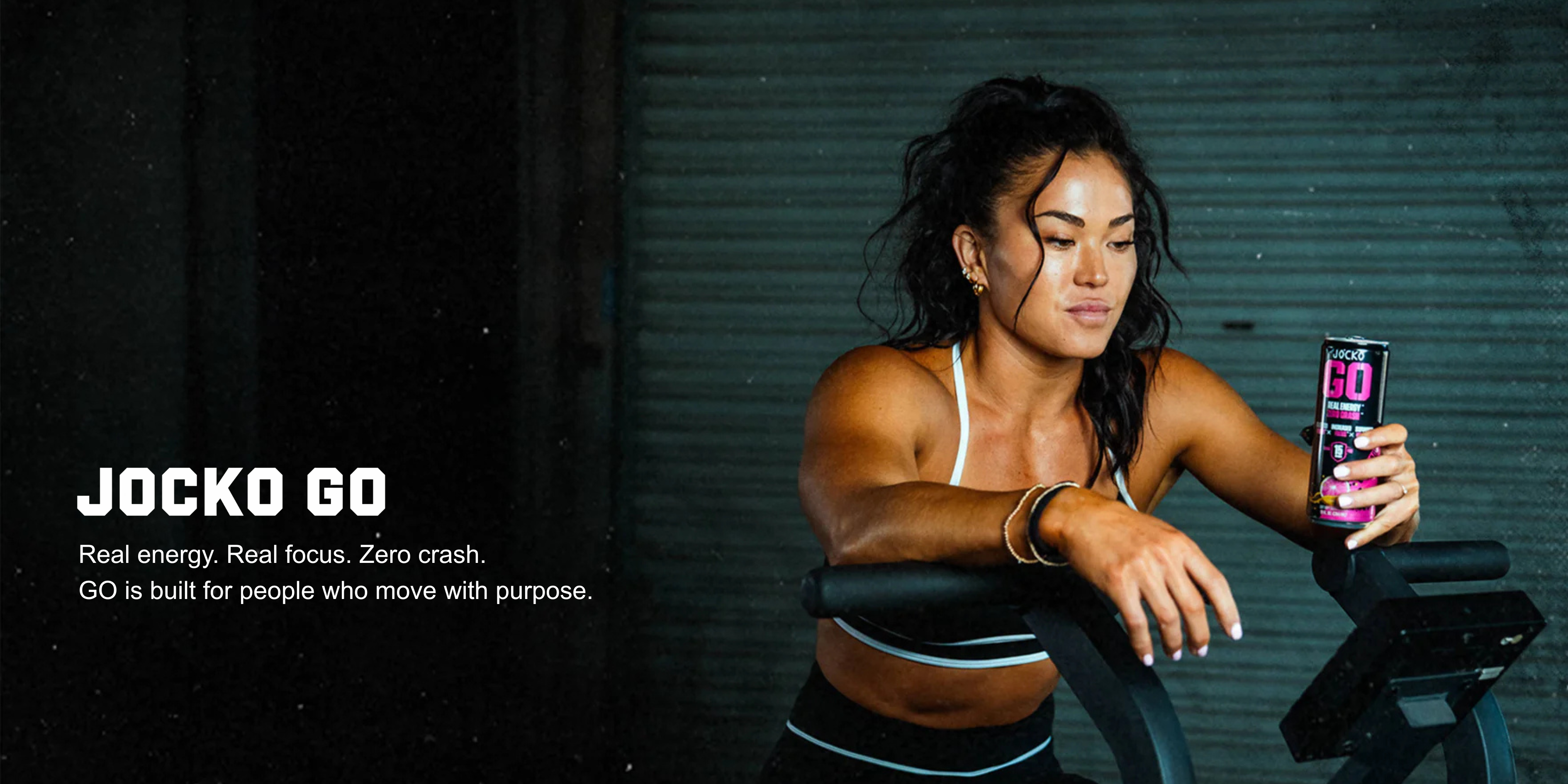 Woman on a stationary bike holding a Jocko Go energy drink can with a dark background.